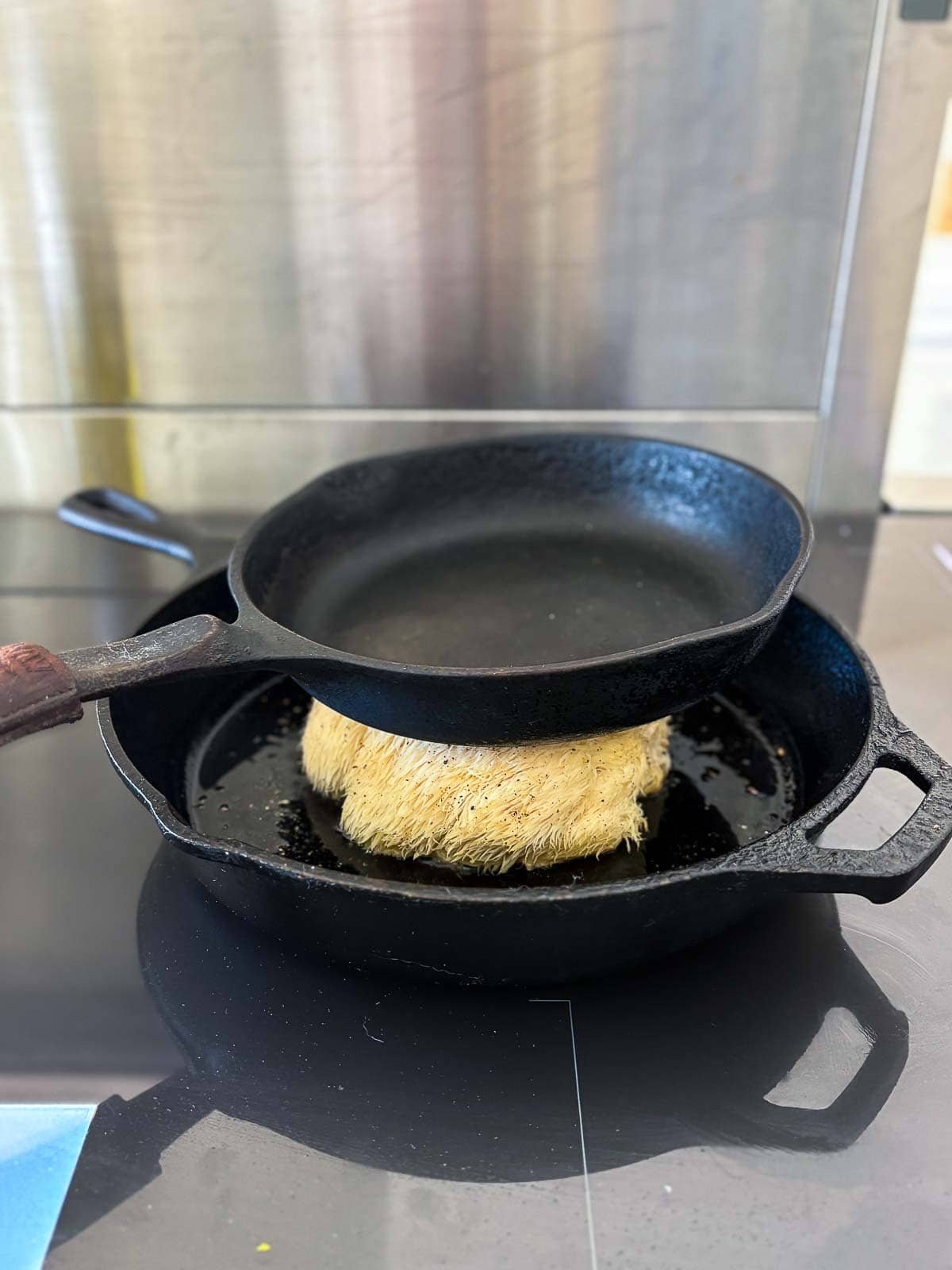 Whole lion's mane mushroom in an oiled skillet with another smaller skillet on top of it pressing it down.